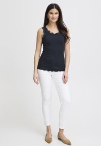 Črni brezrokavnik z nazobčanim čipkastim izrezom, združen s prilagojenimi belimi hlačami in bež balerinkami. Gladek material, krojen dizajn.