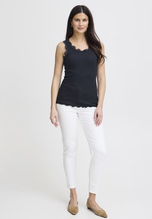 Top negru fără mâneci cu decolteu din dantelă cu margini ondulate, asortat cu pantaloni albi mulați și pantofi bej fără toc. Material neted, croială ajustată.