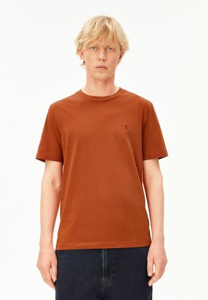 Blonder Mann mit kurzen Haaren trägt ein schlichtes braunes T-Shirt mit kurzen Ärmeln und dunkle Jeans, steht vor weißem Hintergrund.