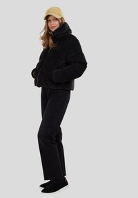 Mustapintainen corduroyn puffer-takki korkealla kauluksella, yhdessä mustien farkkujen ja mustien tennareiden kanssa. Vaalea lippalakki päässä.