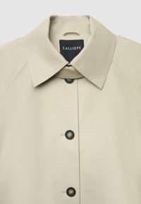 Beige Jacke mit Kragen und glatter Textur, mit schwarzen Knöpfen und einem Etikett mit der Aufschrift "CALLIOPE" am inneren Kragen.