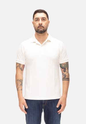 Hombre con barba y tatuajes que lleva una camiseta tipo polo blanca lisa de manga corta y vaqueros azules, de pie frente a un fondo neutro.