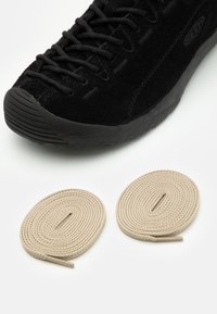 Czarne zamszowe sneakersy z teksturowanym przodem, z okrągłą gumową podeszwą. Dwa spiralne beżowe sznurowadła umieszczone poniżej buta.