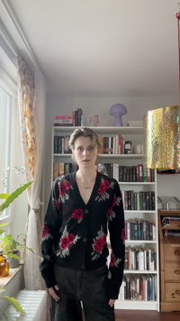 Mustat kardiganit punaisella ruusupistolla, pyöreillä napilla ja pehmeällä tekstuurilla, yhdistetty tummiin housuihin. Taustalla on kirjahyllyjä ja sisustusta.