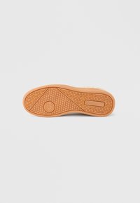 Καφέ ανοιχτό πέλμα παπουτσιού με υφή από κυκλικά και τετραγωνισμένα σχέδια, παρουσιασμένο πάνω σε απλό γκρι ανοιχτό φόντο.