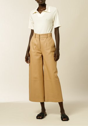 Osoba ubrana w biały top z krótkim rękawem i kołnierzykiem, wysokie spodnie o szerokich nogawkach w kolorze beżowym oraz czarne sandały typu slide z odkrytymi palcami, stojąca na neutralnym tle.