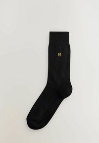 Sort bomuldsok med ribbet manchet, prydet med et lille gult logo i det øverste venstre hjørne. Glat tekstur og klassisk design.