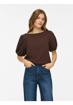 Femme aux cheveux bruns ondulés portant un haut marron foncé à manches bouffantes et un jean bleu taille haute, les mains dans les poches sur fond blanc.