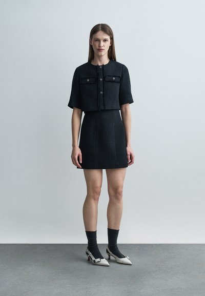 Γυναίκα που στέκεται μπροστά σε απλό φόντο, φοράει μαύρο κοντό μπουφάν με κοντά μανίκια και φούστα ταιριαστή, με μαύρες κάλτσες και λευκά τακούνια.