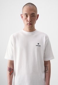 T-shirt en coton blanc à manches courtes, avec un petit logo noir "snow peak" sur la poitrine. Tatouages colorés visibles sur le bras.