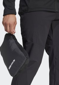 Svart isolert jakke pakket i en kompakt, svart nettingpose. Hånden holder posen, og viser frem materialet og det strømlinjeformede designet.