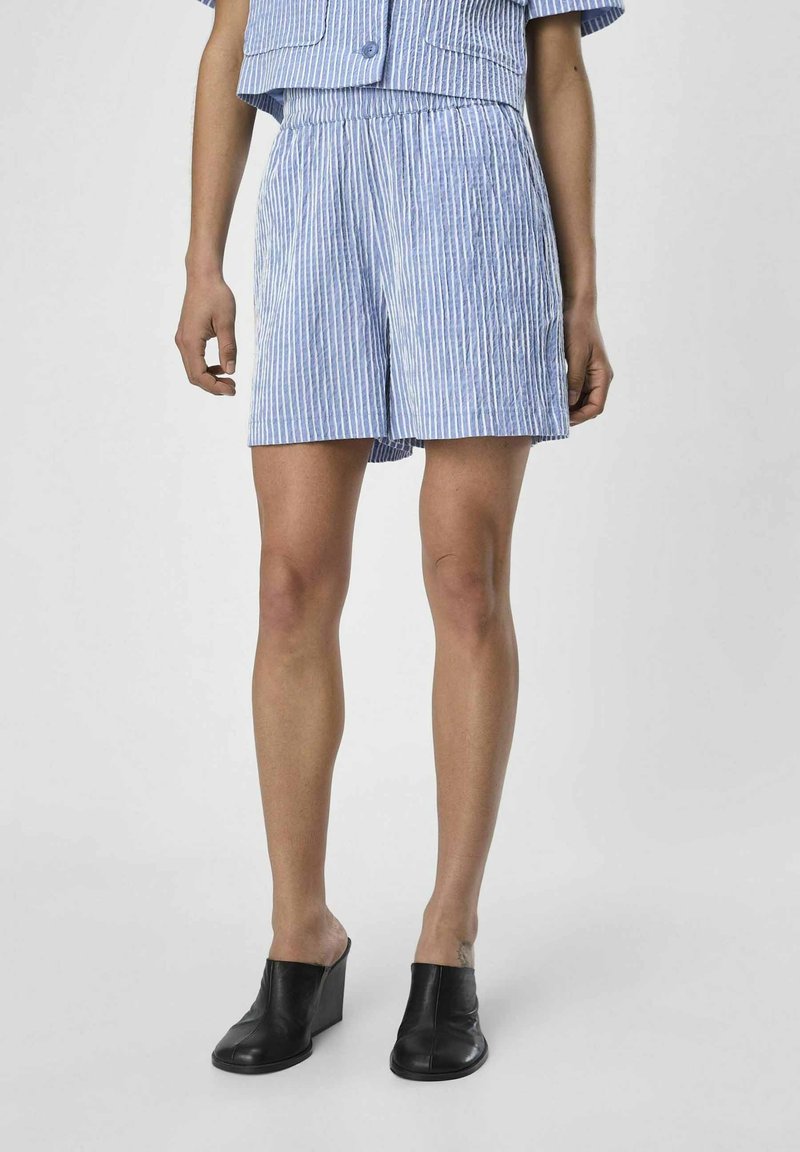 Personne portant un short à rayures verticales bleu clair et blanc et une chemise courte assortie, associée à des chaussures compensées noires, debout sur un fond uni.