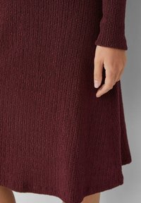 Bordeauxfarbendes, strukturiertes Kleid mit Rippenmuster; verfügt über lange Ärmel und einen ausgestellten Saum. Hand teilweise auf dem Stoff sichtbar.