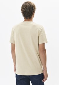 T-shirt beige à manches courtes en tissu doux, avec un col rond et une coupe décontractée, affichant un dos uni. Porté avec un jean bleu foncé.