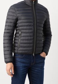 Giacca nera imbottita con colletto alto, caratterizzata da linee trapuntate orizzontali, tasche laterali con zip e chiusura frontale con zip. Abbinata a jeans blu.