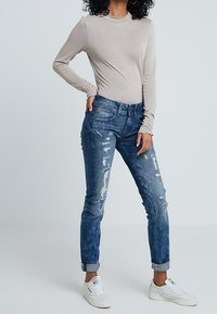 Top beige de manga larga combinado con jeans de mezclilla azul desgastados. Los jeans tienen desgastes y un ajuste relajado, junto con zapatillas blancas.