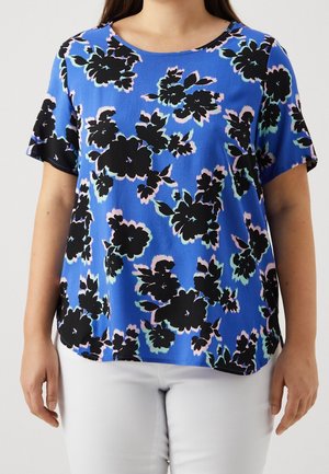 Vrouw draagt een blauw shirt met korte mouwen met zwart bloemmotief en witte broek, staand voor een eenvoudige lichte achtergrond.