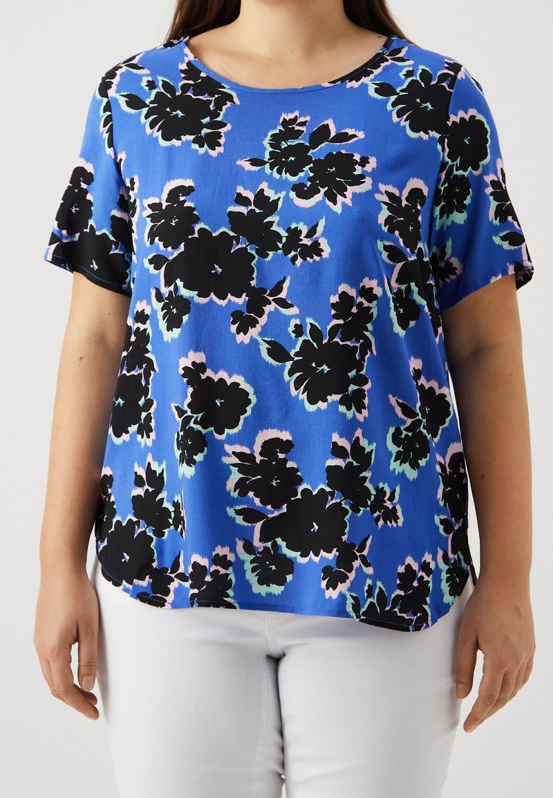 Femme portant un haut bleu à manches courtes avec un motif floral noir et un pantalon blanc, debout devant un fond clair uni.