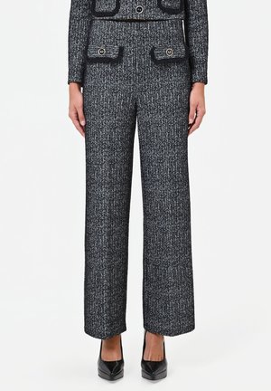 Femme portant un pantalon large en tweed noir et blanc avec une veste assortie et des escarpins noirs pointus sur fond blanc.