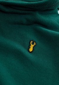 Un tissu vert au toucher lisse présente un logo de cerf brodé en jaune, bordé de noir, situé sur le côté gauche.