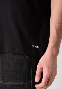 Černé bavlněné tričko s kulatým výstřihem a krátkými rukávy, na dolním levém lemu s bílým logem "HUGO". Nosit s tmavými džíny.