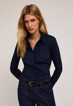 Marineblauwe blouse met kraag, gemaakt van gladde stof, met lange mouwen en een knoopsluiting aan de voorkant, gecombineerd met denim broek met kralenaccenten.