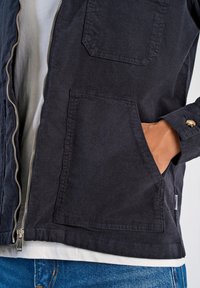 Granatowa kurtka z płótna z zamkiem, dwoma dużymi kieszeniami z przodu i mankietami zapinanymi na guzik. Ma teksturowane wykończenie i jest noszona na białej koszuli.