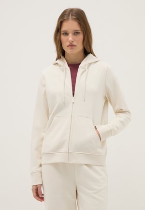 Junge Frau trägt einen cremefarbenen Kapuzenpullover mit Reißverschluss und Kordeln und passende Hose, steht vor einem einfarbigen hellen Hintergrund.