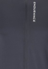 Marineblaues Sportoberteil mit glatter Textur, das mit weißen „ENDURANCE“-Buchstaben sowie einem zentralen Reißverschluss ausgestattet ist. Schlichtes, tailliertes Design.