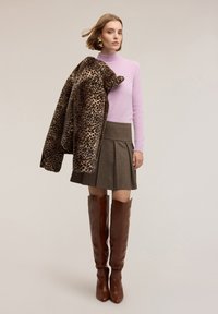 Jasnoróżowy sweter, beżowa spódnica w kratę, brązowe kozaki za kolano oraz płaszcz w panterkę zarzucony na ramię. Proste tło.