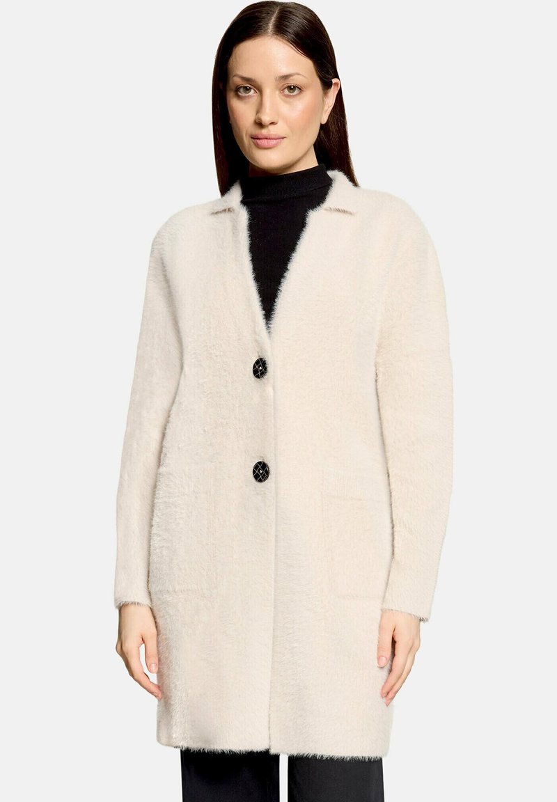 Femme portant un long manteau texturé crème avec deux boutons noirs sur un col roulé noir, debout devant un fond clair uni.