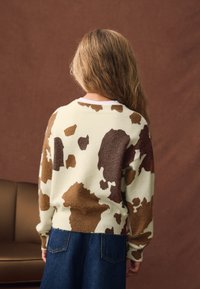 Krem pulover z rjavim in temnobež vzorcem kravje dlake, z zaobljenim izrezom in rebrastimi manšetami, kombiniran s temnimi denim kavbojkami.