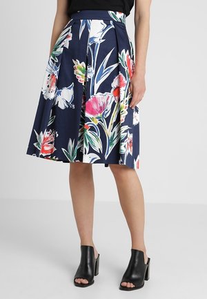 Kvinde iført navy plisseret nederdel med stort farverigt blomsterprint og sorte åbne blockhæle, stående mod en ensfarvet baggrund.