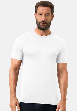 Mann mit Bart, der ein schlichtes weißes T-Shirt mit kurzen Ärmeln trägt und vor einem hellgrauen Hintergrund steht.
