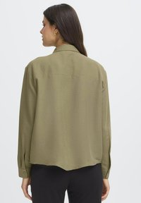 Donna con lunghi capelli scuri che indossa una maglietta a maniche lunghe verde oliva e pantaloni neri, ripresa di spalle su uno sfondo neutro.