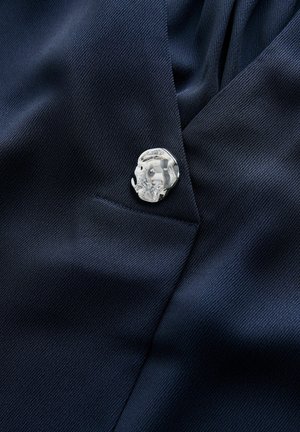 Tmavě modrá texturovaná tkanina s lesklým stříbrným knoflíkem nepravidelného tvaru poblíž švu nebo okraje kapsy.