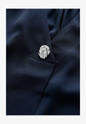 Țesătură texturată albastru închis cu o nasture argintiu lucios, cu formă neregulată, aproape de o cusătură sau marginea unei buzunare.