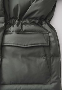 Veste matelassée vert foncé avec taille cintrée, une grande poche à rabat et une texture lisse et brillante.