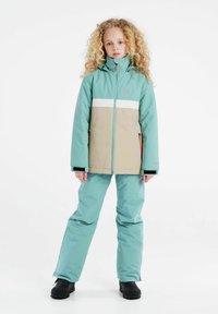 Een turquoise en beige winterjack met een witte horizontale strook, ritszakken en bijpassende turquoise broek. Zwarte schoenen maken de outfit af.