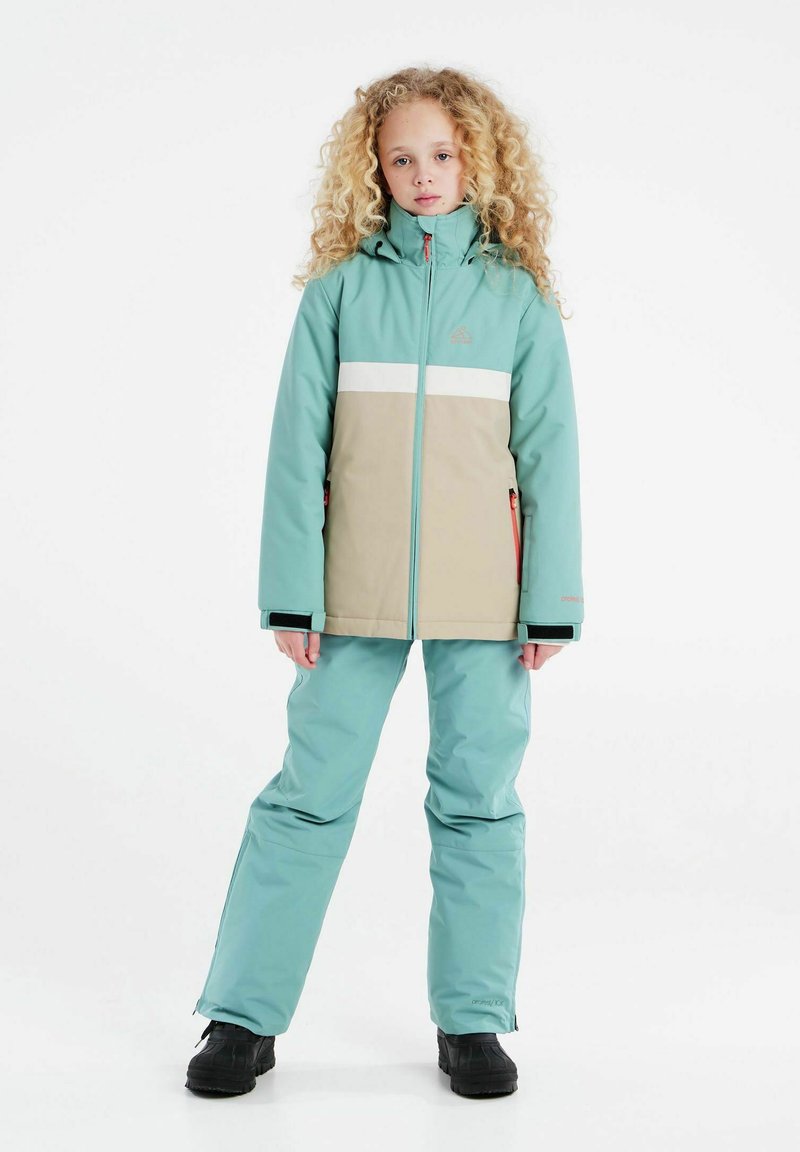 Een turquoise en beige winterjack met een witte horizontale strook, ritszakken en bijpassende turquoise broek. Zwarte schoenen maken de outfit af.