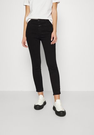 Mujer que lleva camiseta blanca, vaqueros negros ajustados y zapatillas blancas con suelas negras gruesas, de pie sobre un suelo gris claro.