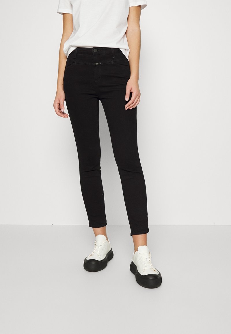 Femme portant un t-shirt blanc, un jean noir slim et des baskets blanches avec des semelles épaisses noires, debout sur un sol gris clair.