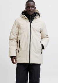 Mustanahuline inimene, kes kannab beežikat kapuutsiga talvejopet musta tõmblukuga ja musti pükse, seisab ühevärvilise heleda tausta ees.