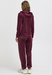 Survêtement en velours bordeaux comprenant un haut à capuche avec une finition texturée et un pantalon élastique assorti, accompagné de baskets blanches imposantes.