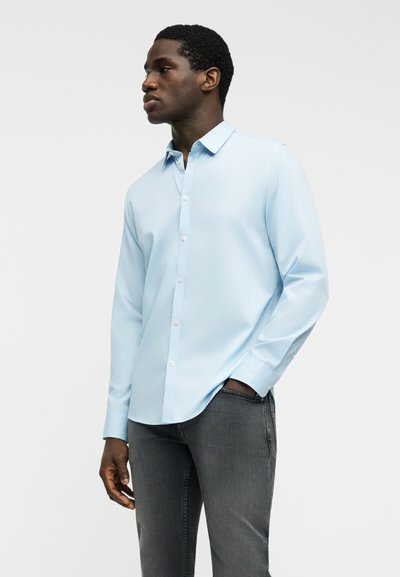 Νέος άνδρας που φοράει ανοιχτό μπλε πουκάμισο με κουμπιά και σκούρο γκρι τζιν, στέκεται με το ένα χέρι στην τσέπη, κοιτάζοντας προς τα αριστερά.