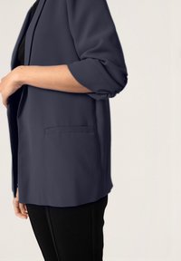 Blazer ligero de color azul marino con mangas enrolladas y un bolsillo en la parte frontal. Llevado sobre una blusa negra, combinado con pantalones negros.