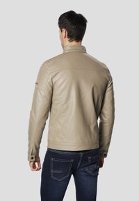 Beżowa skórzana kurtka z pikowanymi akcentami na ramionach, usztywnionym kołnierzem i zapinanymi na guziki mankietami, noszona na ciemnoniebieskich dżinsach. Czysty, nowoczesny design.