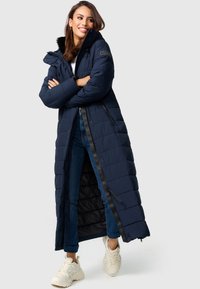 Marinblå puffjacka med lång design, quiltad textur, foder av päls vid huvan, framdragkedja och svarta accenter, i kombination med blå jeans och vita sneakers.