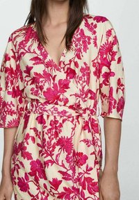 Robe portefeuille à motifs floraux avec un décolleté en V, des manches volantes et de vives fleurs roses sur un fond beige. Détail de taille nouée.