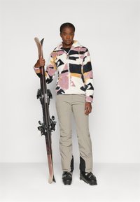 Flisast jakna z rožnato, črno in rumeno abstraktno vzorcem, v kombinaciji z bež smučarskimi hlačami. Smuči z vezmi, držane v levi roki.